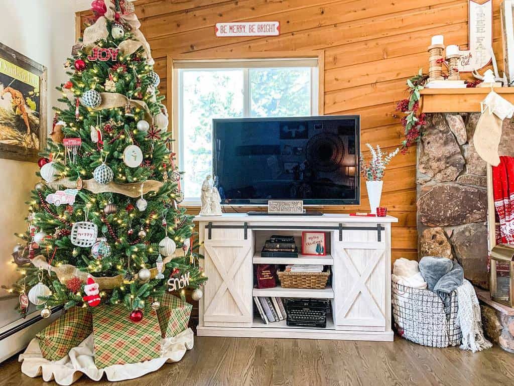 A decorated Christmas tree and stockings adorn a cozy living room with a TV on a white stand, rustic wooden walls, and stone fireplace