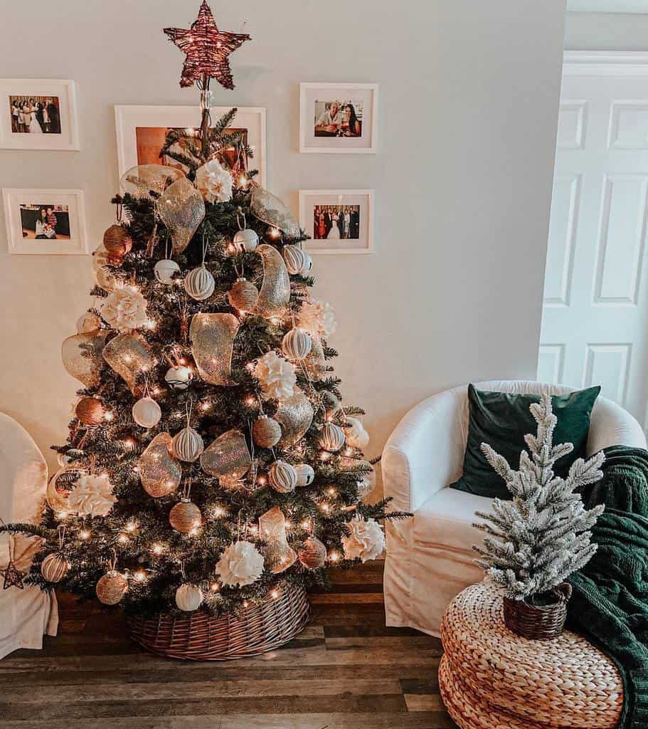 A decorated Christmas tree with ornaments and lights next to two white chairs and a small frosted tree on a wicker stool