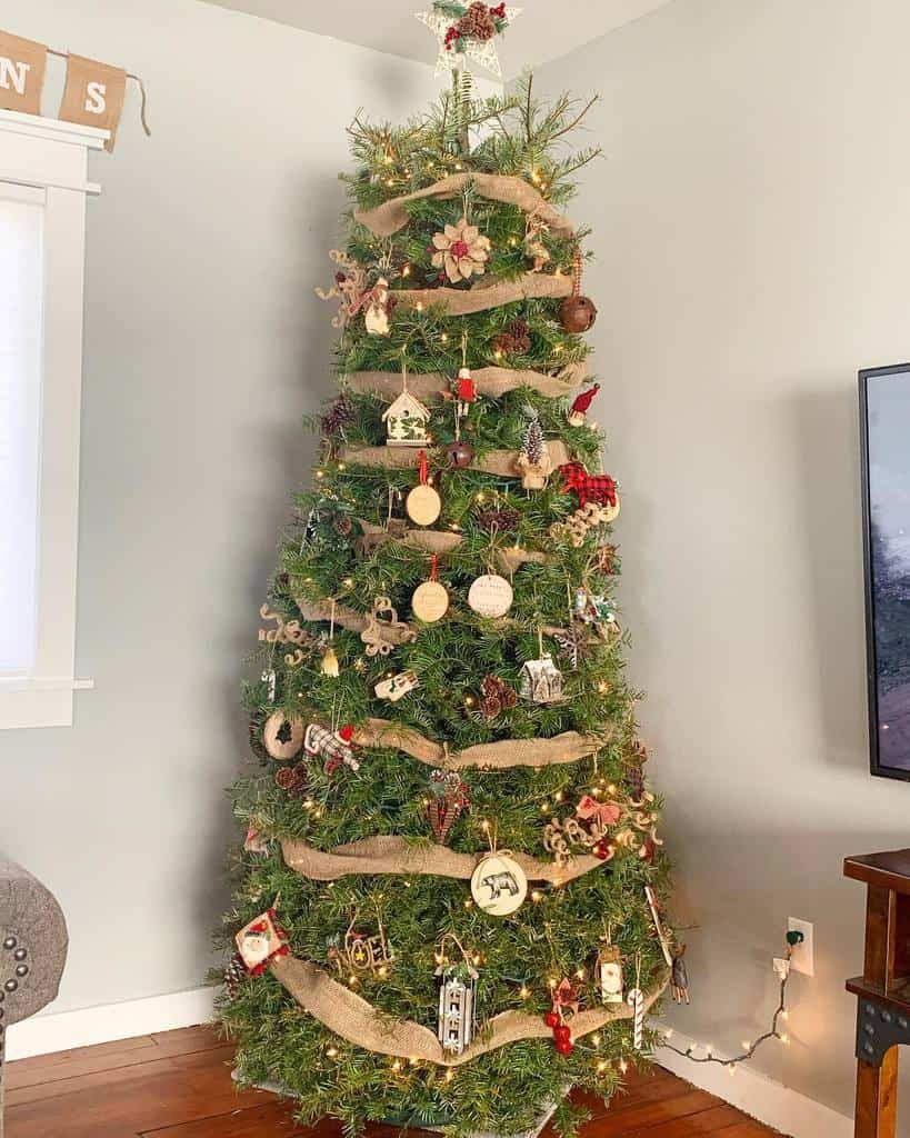 A decorated Christmas tree with ornaments and burlap garland stands in a living room with hardwood floors and a TV nearby