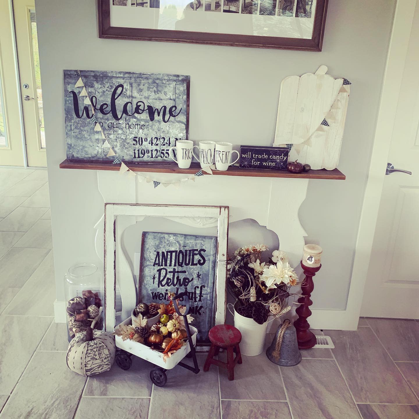 Rustic decor display with welcome signs, jars, flower vases, and a mini cart filled with small pumpkins on a tiled floor