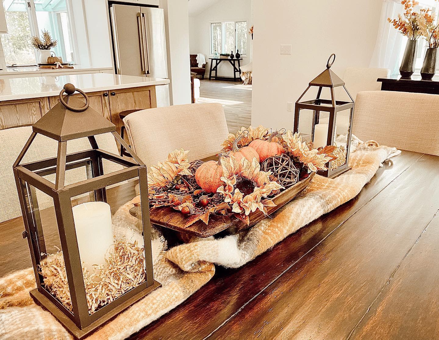 A fall-themed dining table with lanterns, candles, pumpkins, and autumn leaves centerpiece on a wooden table with beige chairs