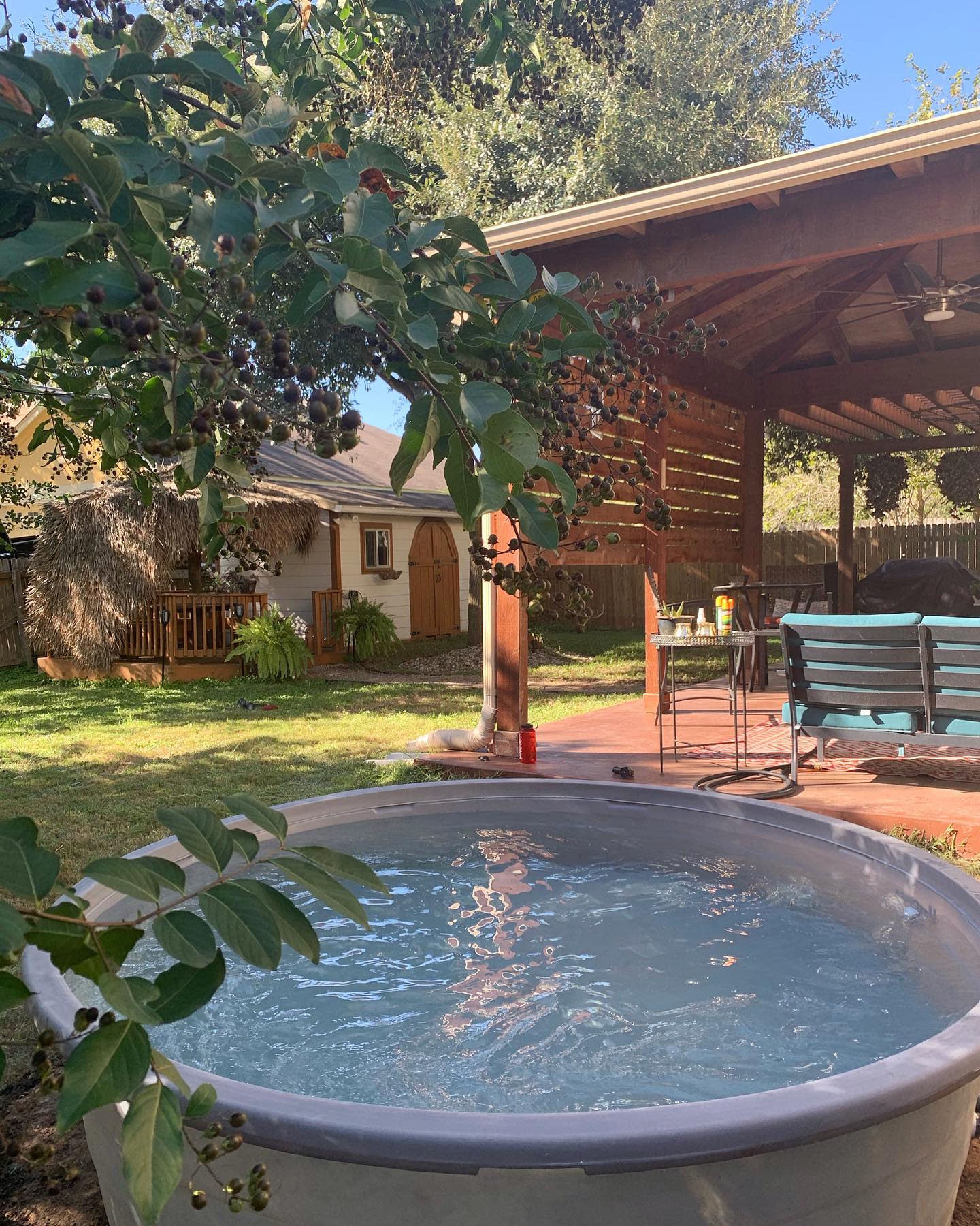 A small round pool in a backyard with a covered patio, seating, and lush greenery, along with a table with drinks in the background