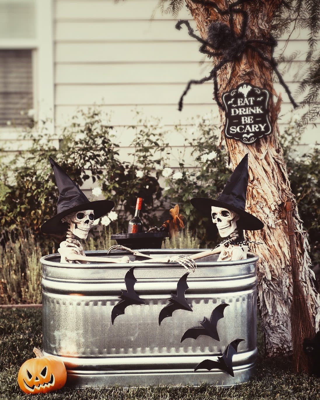 Two skeletons in witch hats relax in a tub with a bottle, surrounded by bats and Halloween decor; a sign reads, "Eat, Drink, Be Scary"