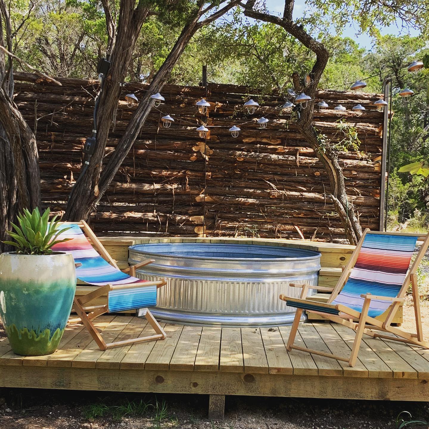 Wooden deck with a small round metal pool, two colorful chairs, a potted plant, and hanging lights in a rustic garden setting