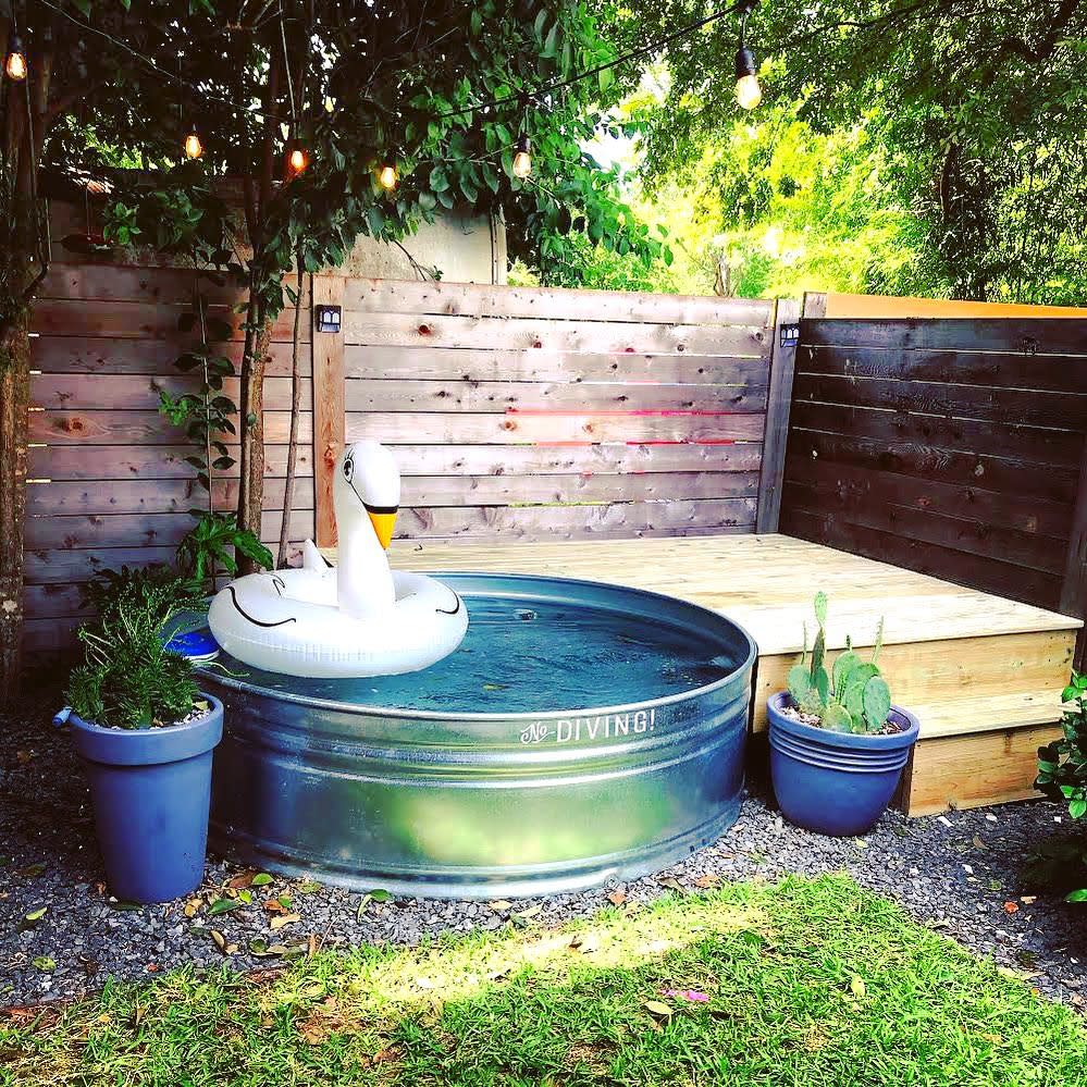 Galvanized metal pool with a swan float in a garden, surrounded by plants and string lights, with wooden deck steps nearby