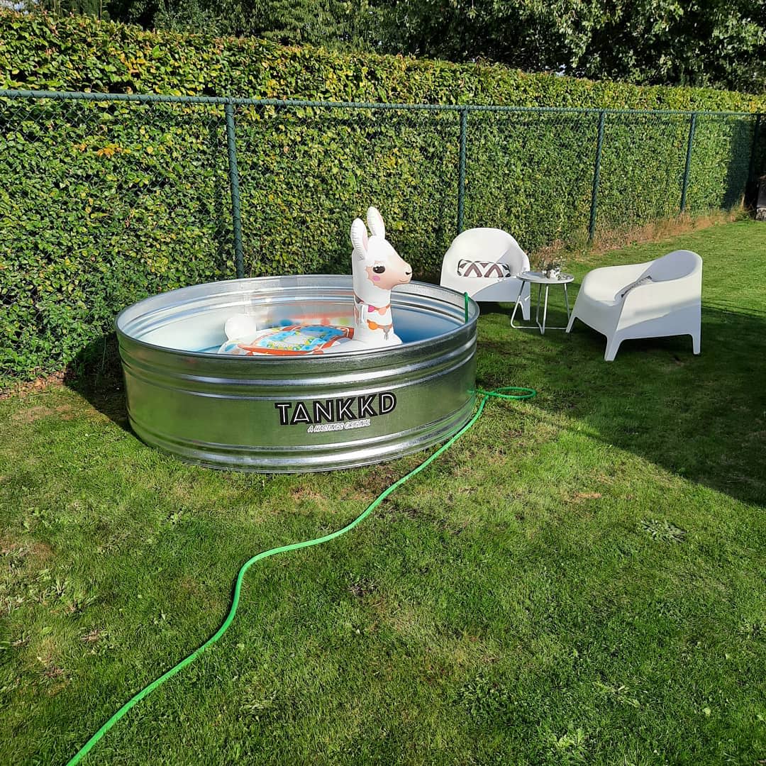 A metal pool with a llama float, surrounded by green grass; two white chairs and a small table are nearby; a hose fills the pool