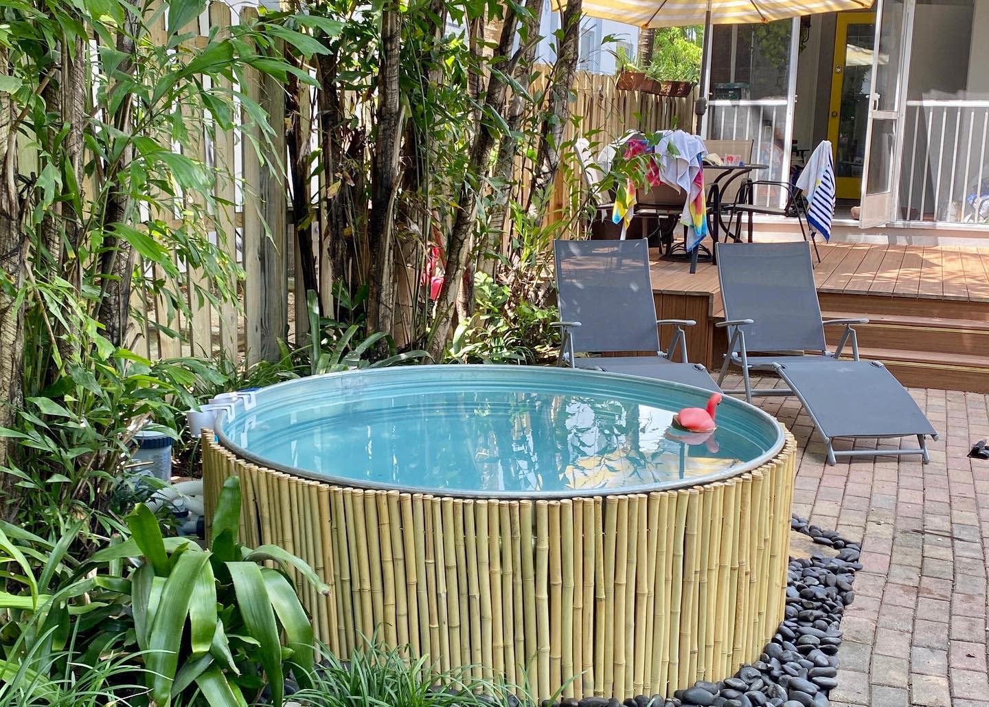 A small, round, bamboo-framed pool surrounded by plants, with two loungers nearby on a patio and towels hanging on a railing in the background