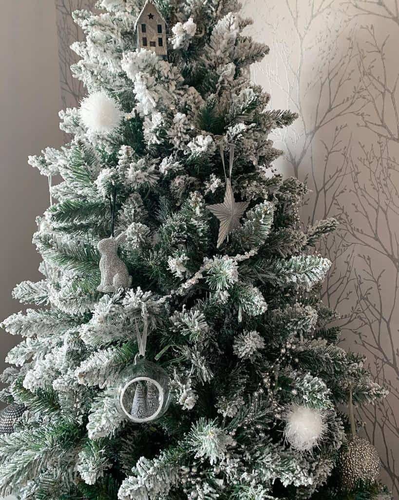 Snowy Christmas tree with white and silver decorations, including a star, house, and pom-poms; wall with tree pattern in background