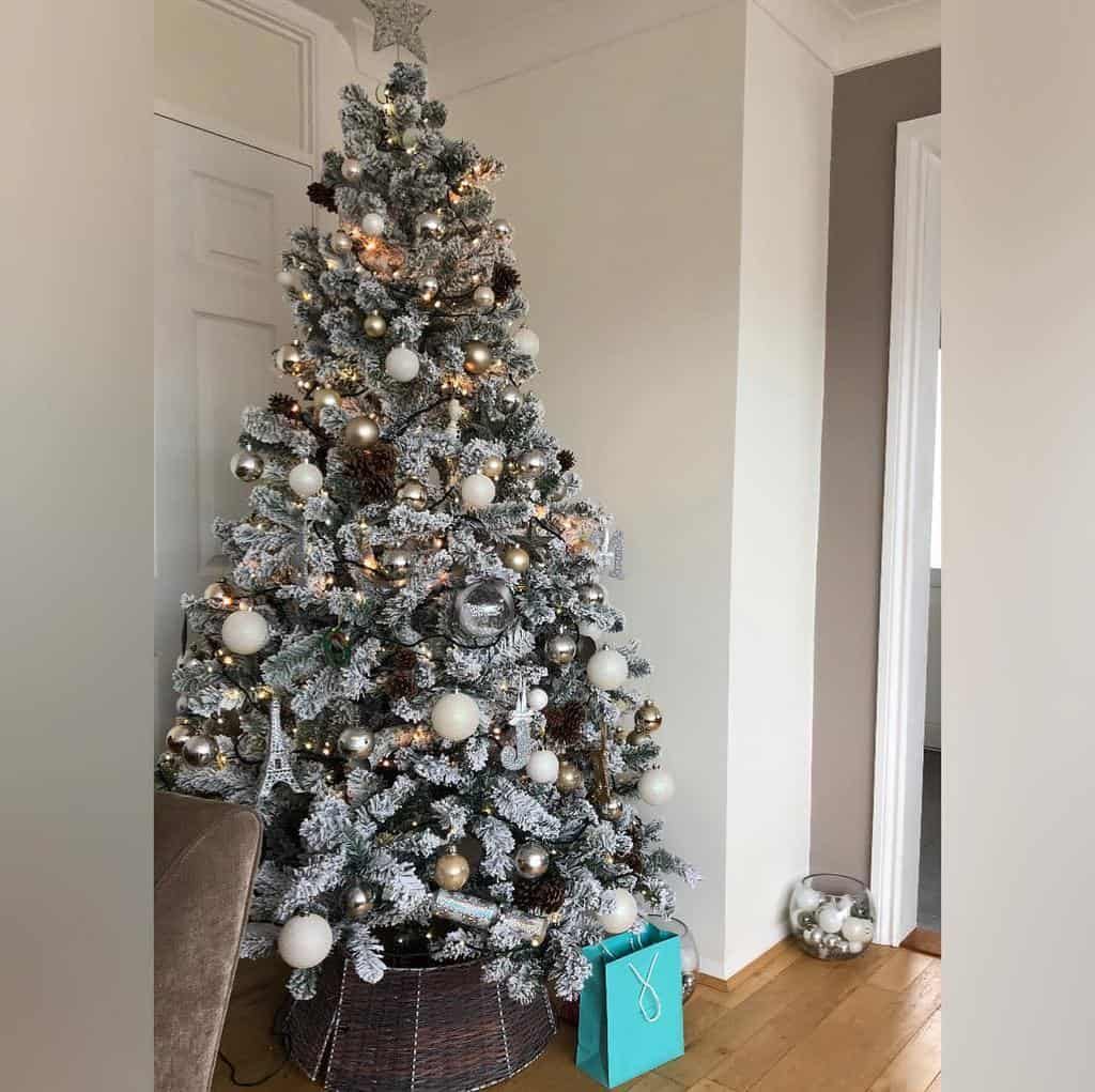 A decorated Christmas tree with white and silver ornaments, warm lights, and a star topper; a small gift bag sits at its base