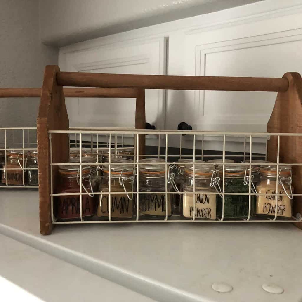 Jars of spices labeled with names, organized in a white wire basket with a wooden handle, sitting on a white shelf