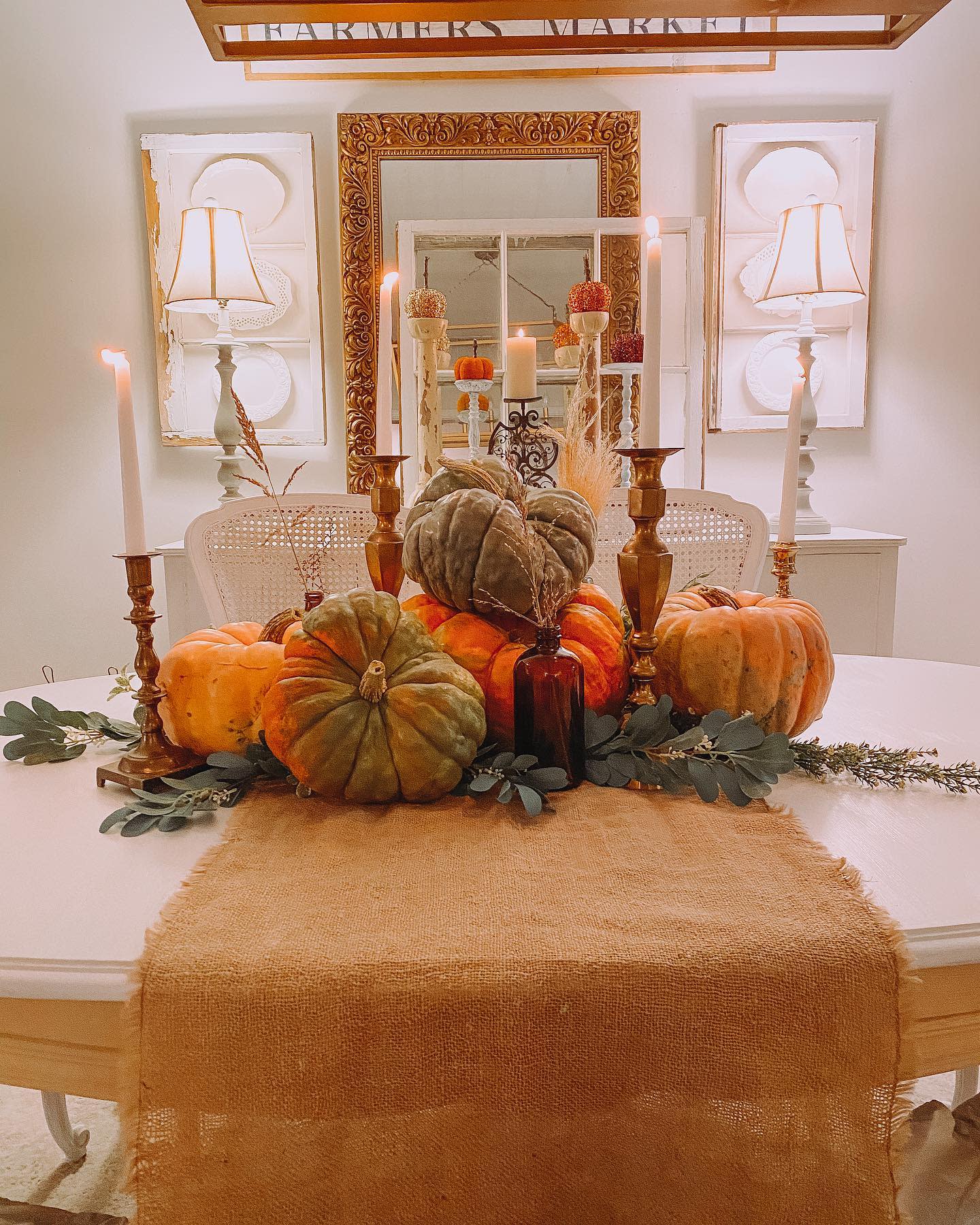 A dining table with a fall centerpiece featuring pumpkins, candles, greenery, and a burlap table runner in a warmly lit room