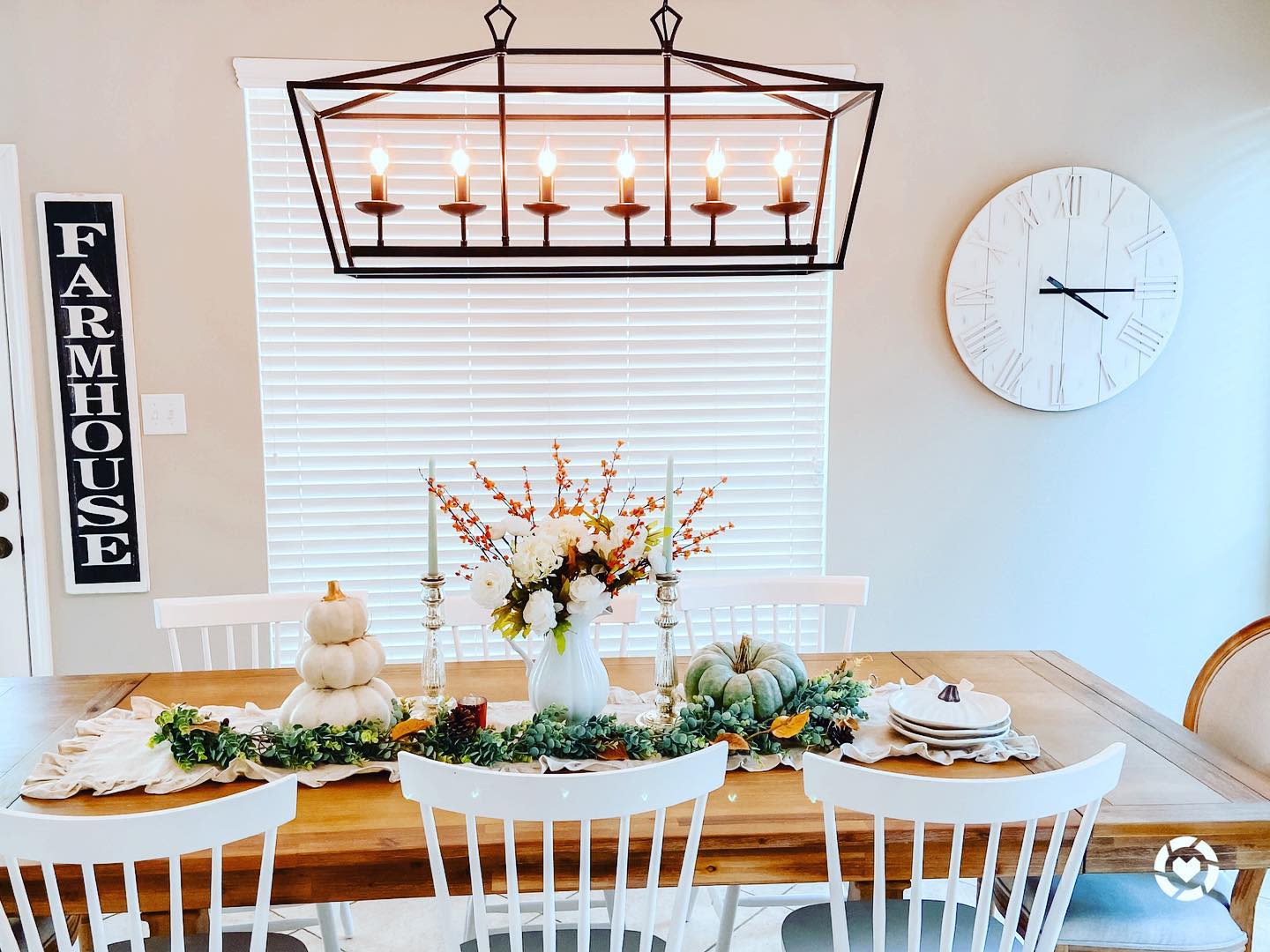 Dining room with a wooden table set for fall, decorated with pumpkins and foliage, a farmhouse sign and clock adorn the walls