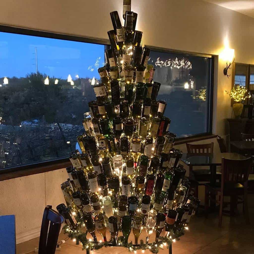 A Christmas tree made of stacked wine bottles, decorated with string lights, stands in a room with large windows