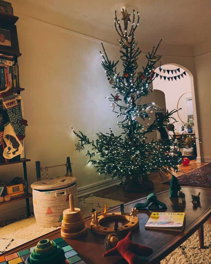 A decorated Christmas tree with lights is in a cozy living room, surrounded by children's toys and a few books on a nearby shelf