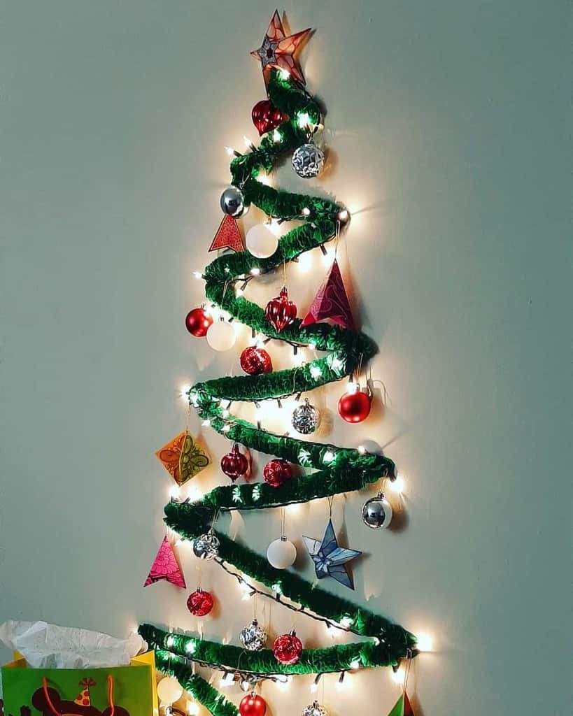 Wall-mounted Christmas tree made of green garland, ornaments, and lights, shaped in a spiral with a star on top