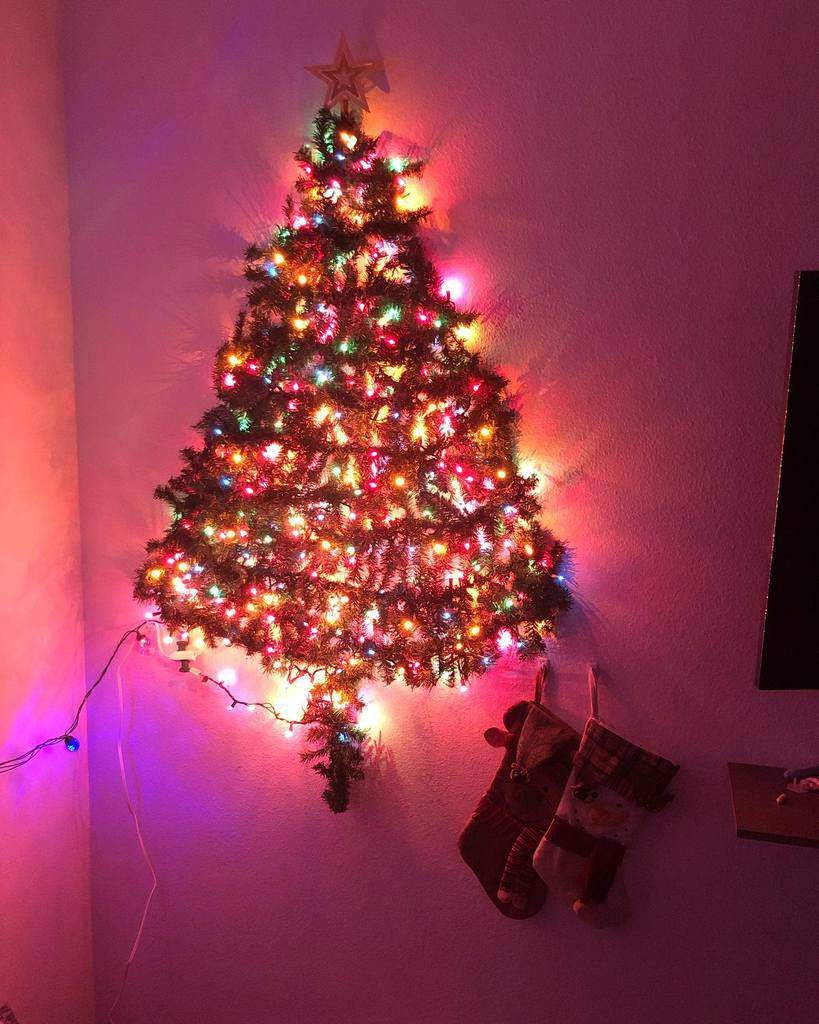 Wall-mounted Christmas tree with colorful lights, topped with a star. Two stockings hang below on the wall