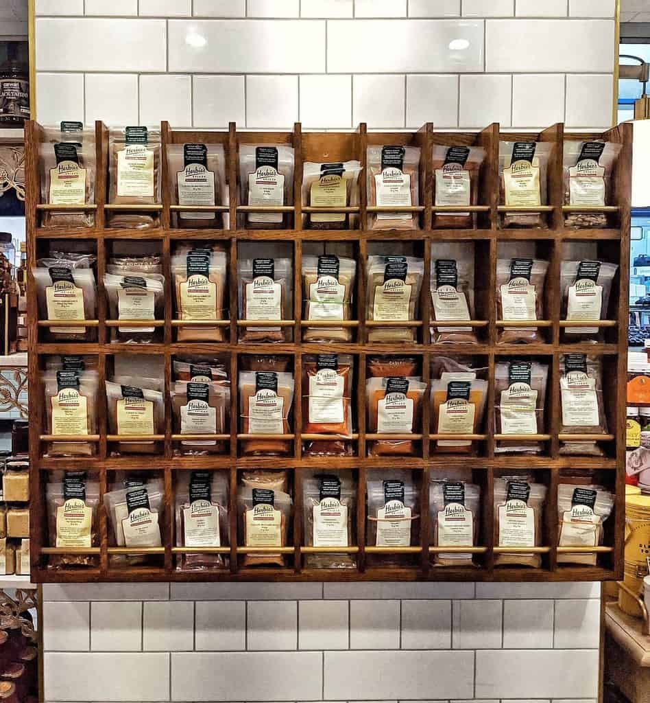 Wall-mounted wooden spice rack with various spices in labeled bags, set against a white tiled background