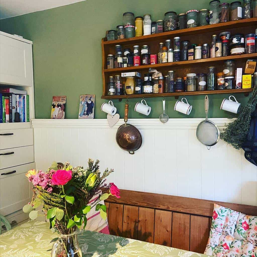 Cozy kitchen corner with a wooden shelf of spices, vintage cookware, a floral bouquet on the table, and books on a white cabinet