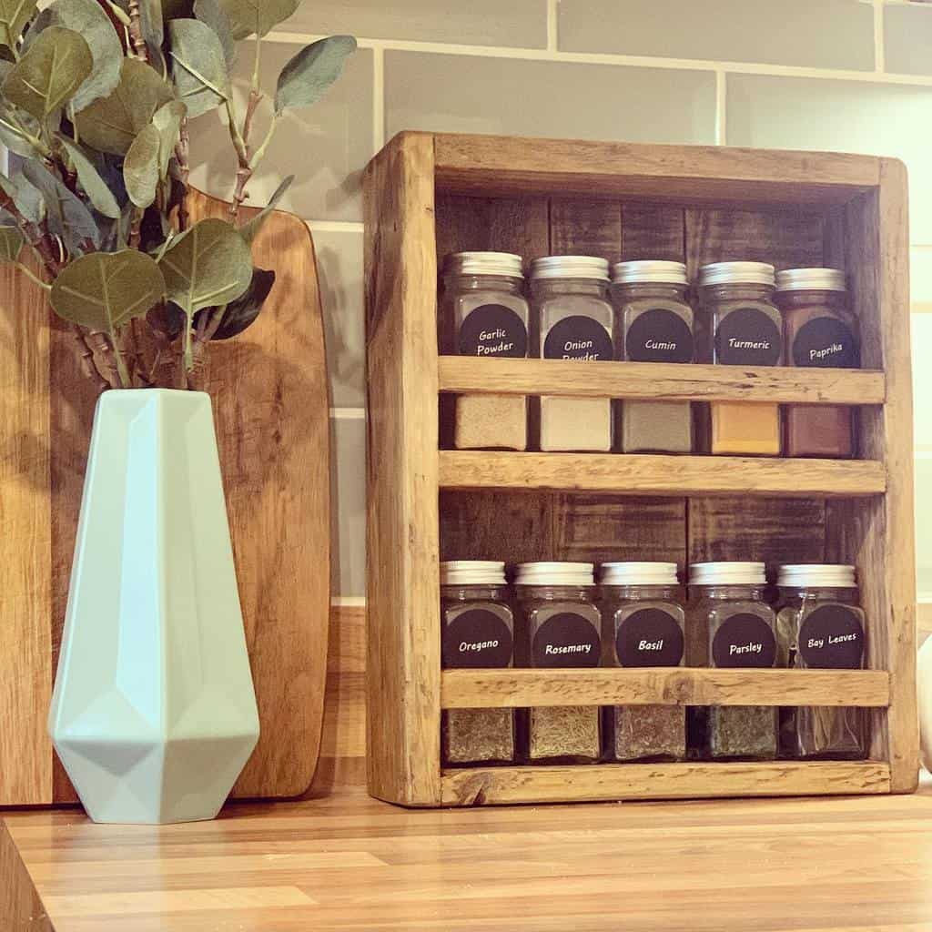 A wooden spice rack with labeled jars, a geometric blue vase with greenery, and a wooden cutting board on a kitchen counter