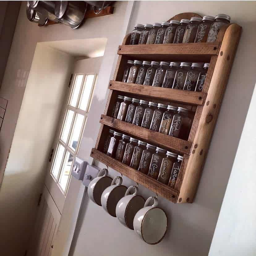 A wall-mounted wooden spice rack with labeled jars and four mugs hanging beneath in a kitchen