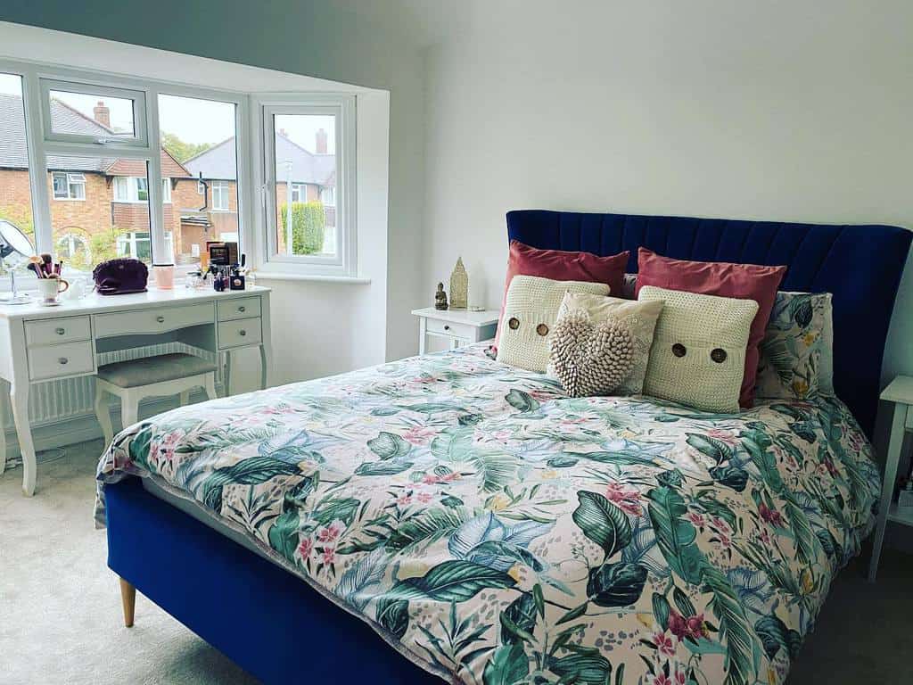 Bright bedroom with a bay window, navy velvet bed, floral bedding, and a vintage-style vanity, creating a cozy and elegant space.