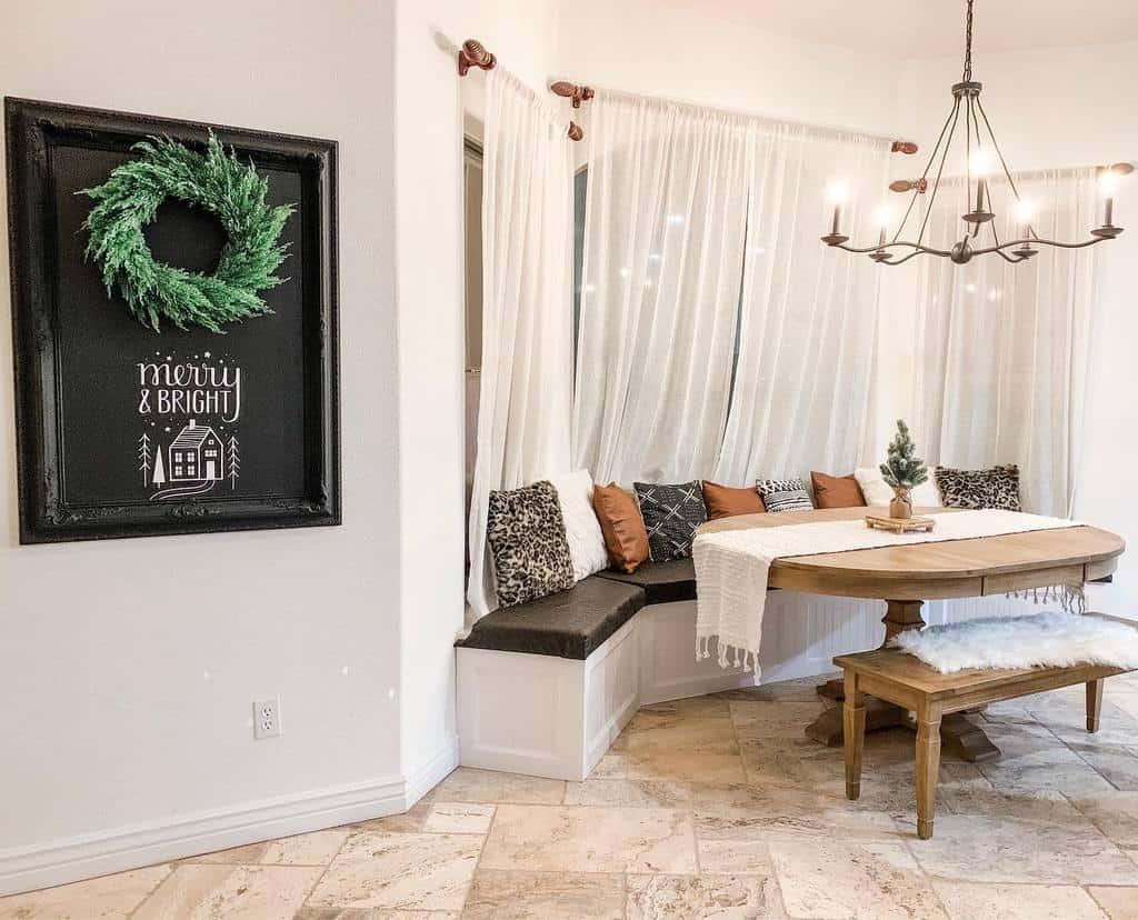 Cozy bay window dining nook with a built-in bench, warm-toned cushions, sheer curtains, and a rustic wooden table for a charming farmhouse feel.