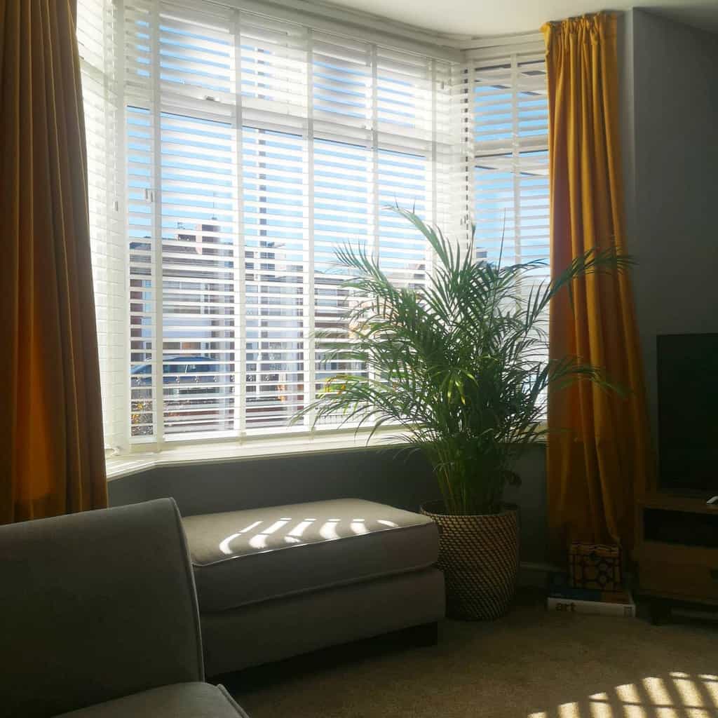 Sunny bay window nook with white blinds, mustard curtains, a cozy bench, and a lush green plant, adding warmth and vibrancy to the space.