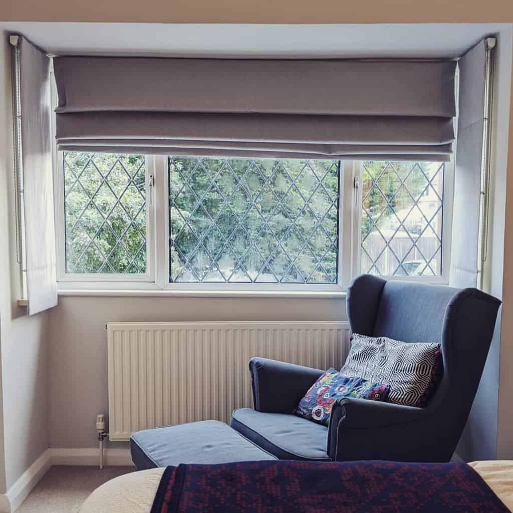 Cozy bay window nook with soft grey Roman blinds, diamond-patterned glass, and a comfy reading chair with a matching footrest.