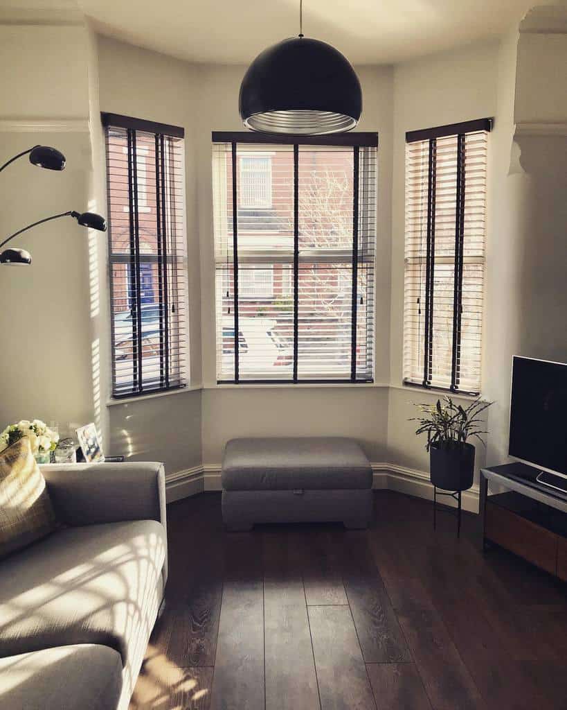 Modern bay window with sleek dark wooden blinds, a cozy ottoman, and stylish lighting, creating a warm and sophisticated living space.