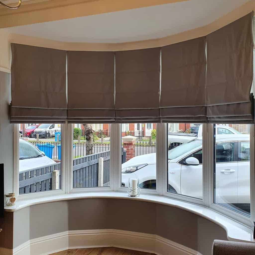 Elegant bay window with tailored gray Roman blinds, soft curved seating, and a bright view, blending privacy with natural light.