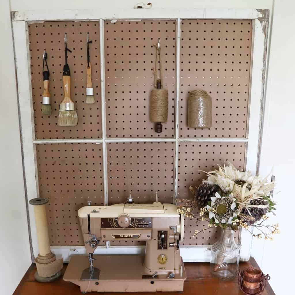 Vintage window repurposed into a pegboard organizer, holding brushes and twine above a retro Singer sewing machine.