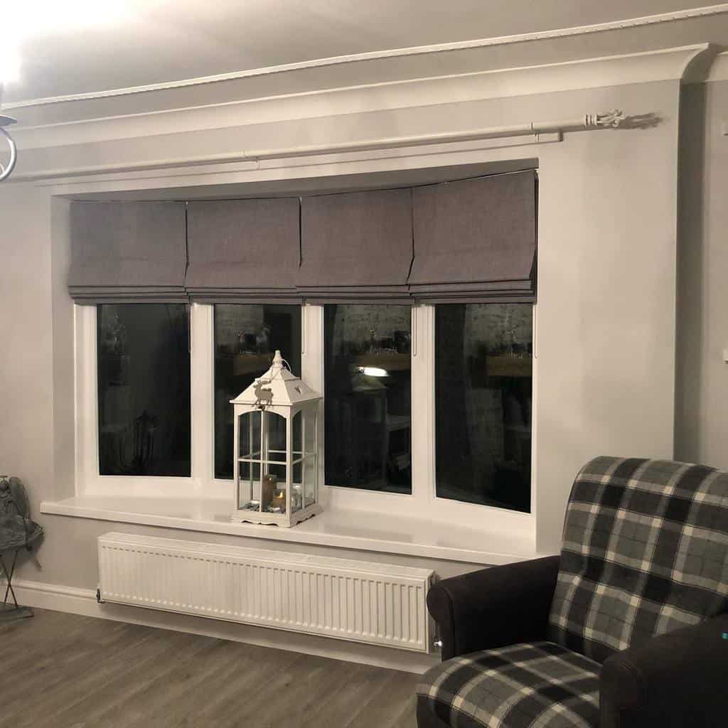 Cozy bay window with gray Roman blinds, a white bench-style sill, and a plaid armchair, creating a warm and inviting living space.