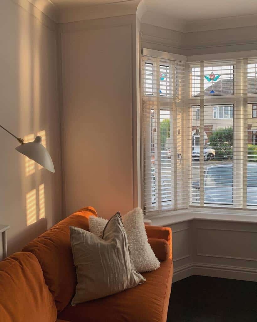 Cozy bay window nook with white blinds, soft stained glass details, and a warm-toned sofa bathed in golden evening light.