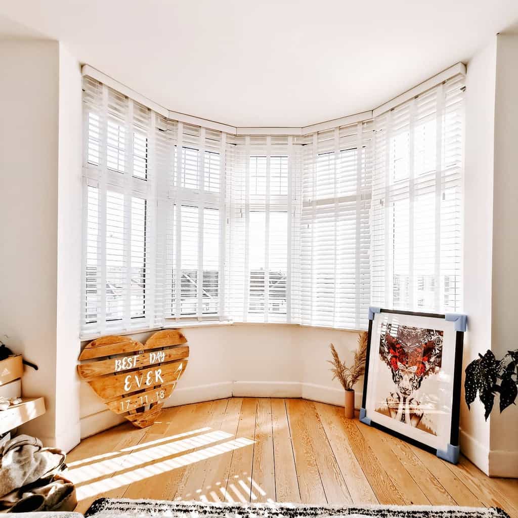 Bright bay window with white wooden blinds, natural light, and warm wood flooring, creating an airy and inviting space with personal decor touches.