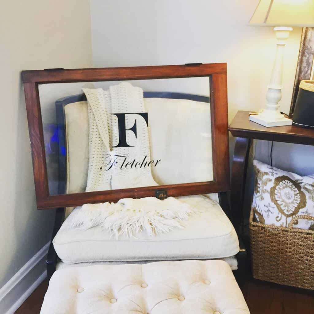 Repurposed old window frame with a personalized monogram design, leaning on a cozy cream-colored armchair in a warm, inviting space.