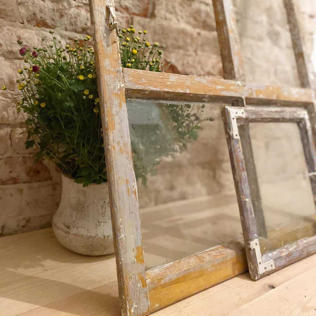 Vintage wooden window frames with peeling paint leaning against a rustic brick wall, with a potted plant in the background.