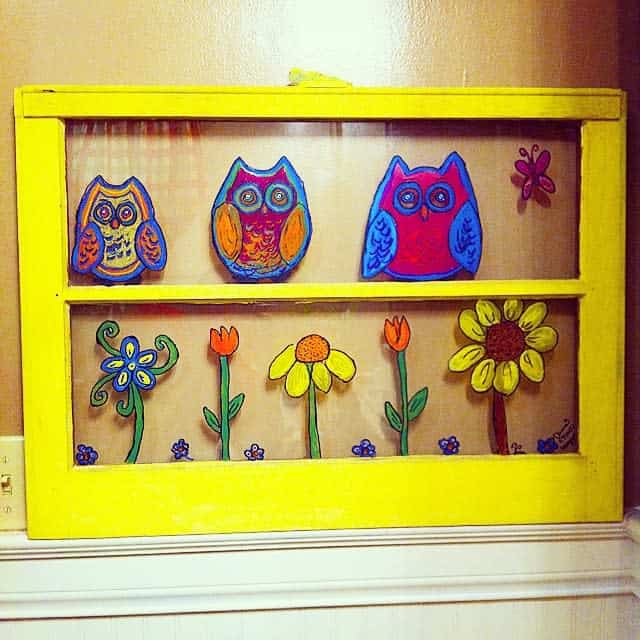 Repurposed old window frame painted bright yellow, featuring hand-painted glass art of colorful owls, flowers, and a butterfly.