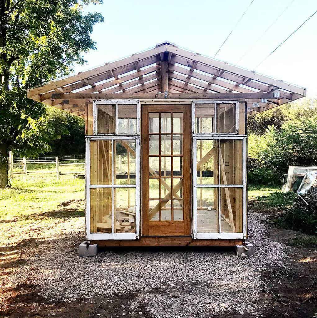 Rustic greenhouse made from repurposed old windows and a wooden door, featuring an open-frame roof in a serene outdoor setting.