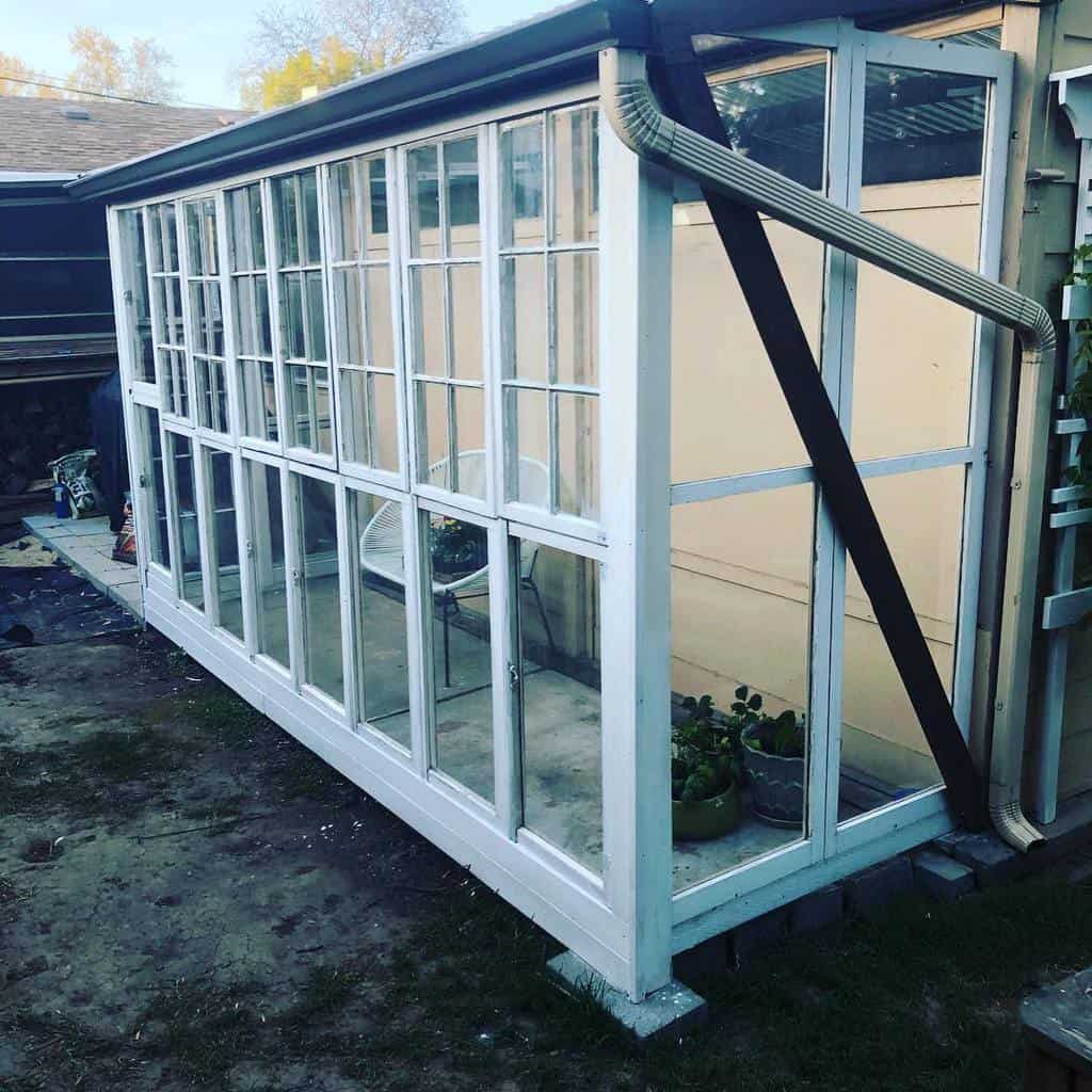 Lean-to greenhouse built from repurposed old windows, attached to a house with a gutter system, creating a cozy space for plants.