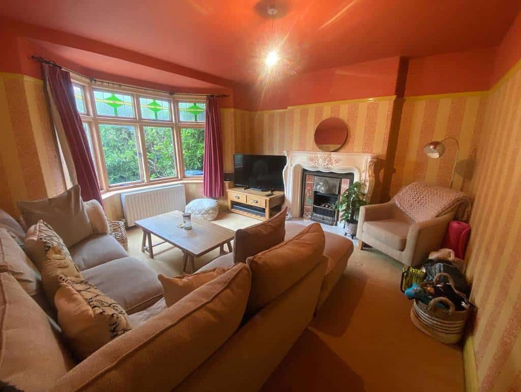 Warm and eclectic living space with a bay window, stained glass details, bold red ceilings, and cozy seating around a classic fireplace.