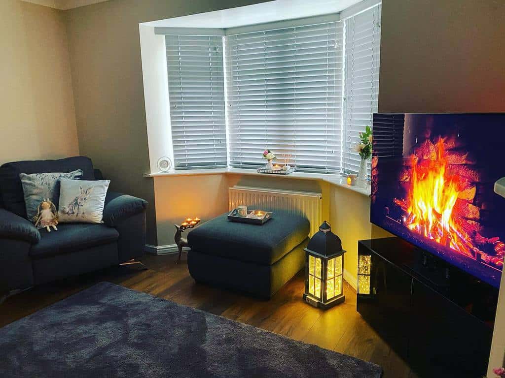 Cozy living room with a bay window, white blinds, soft lighting, and a virtual fireplace, creating a warm and inviting ambiance.