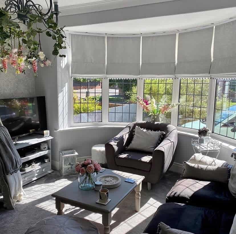 Bright and elegant living room with a curved bay window, soft Roman blinds, cozy seating, and floral accents, creating a charming retreat.