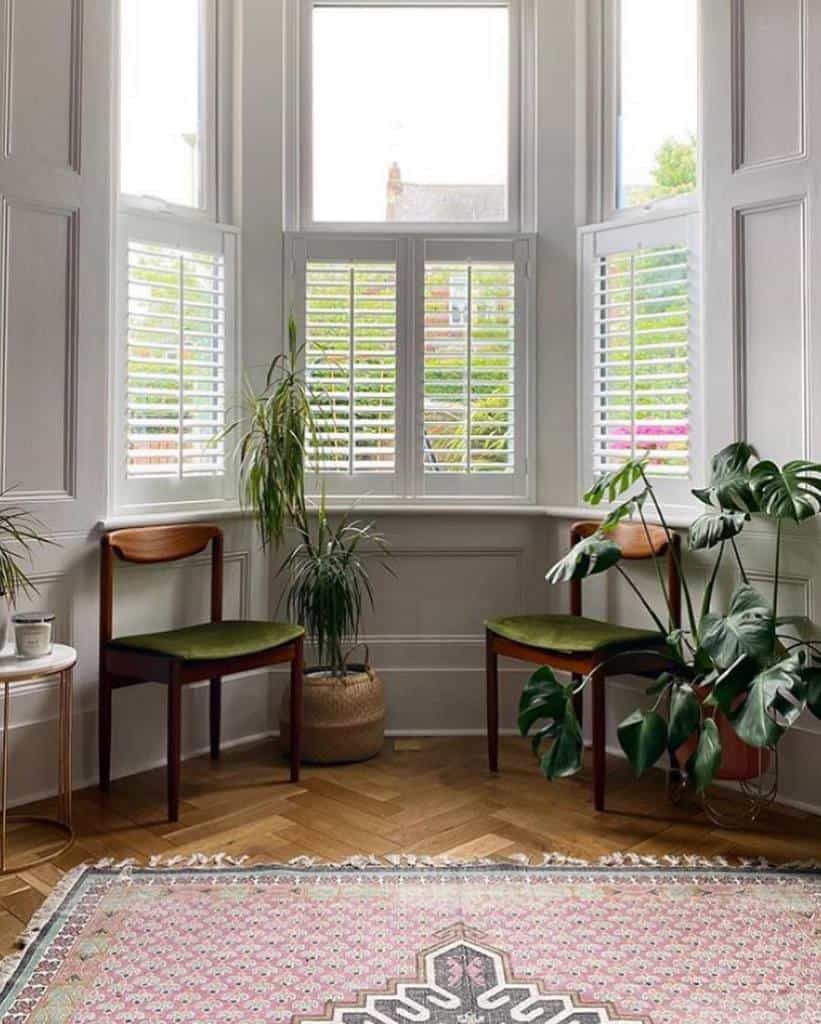 Stylish bay window nook with white shutters, mid-century chairs, lush greenery, and a vintage rug, creating a cozy and elegant space.