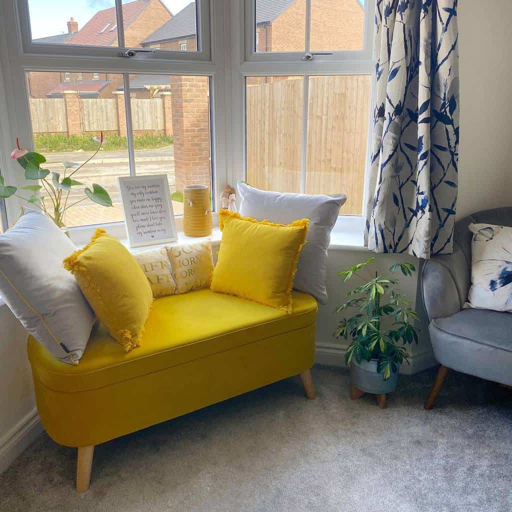 Cheerful bay window nook with a mustard yellow bench, cozy pillows, soft curtains, and greenery, creating a bright and inviting space.