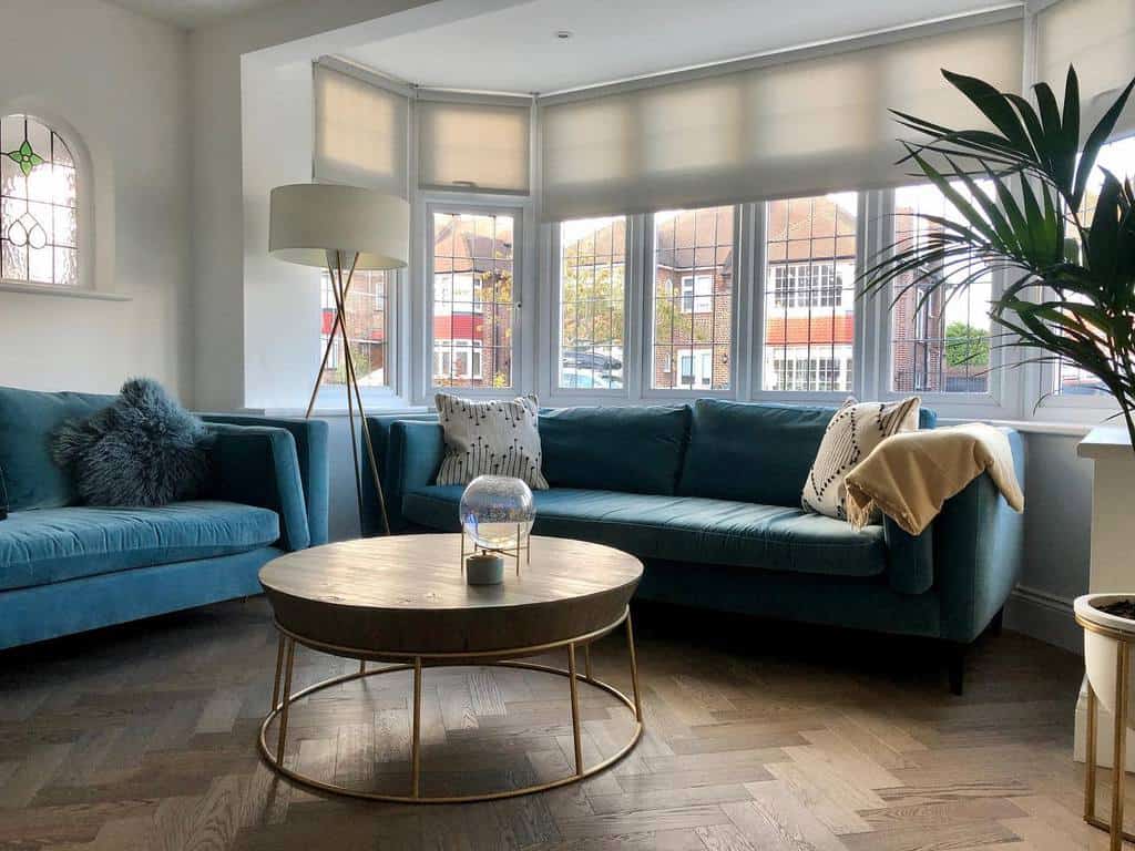 Modern living room with a bay window, soft roller blinds, plush blue sofas, and a chic gold coffee table, creating a stylish and cozy space.