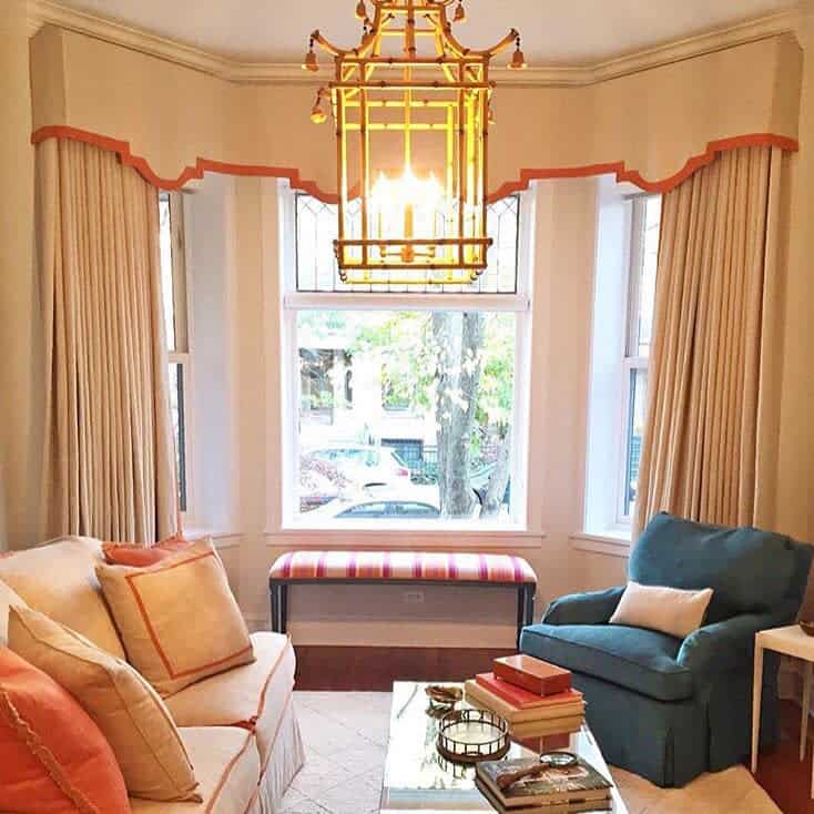 Elegant bay window living room with classic drapes, a statement chandelier, cozy seating, and a striped bench, creating a warm and inviting space.