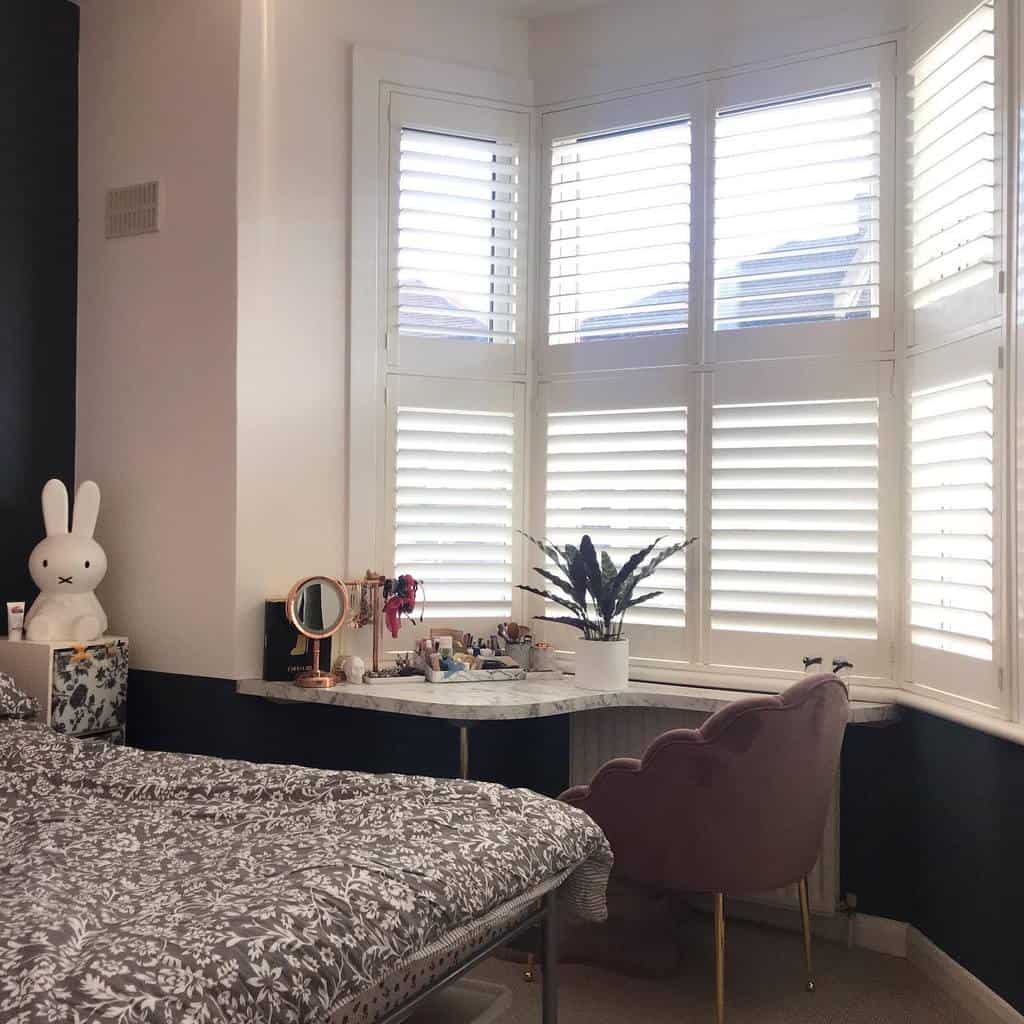 Stylish bay window nook with white plantation shutters, a marble vanity, and a plush pink chair, creating a chic and functional bedroom space.