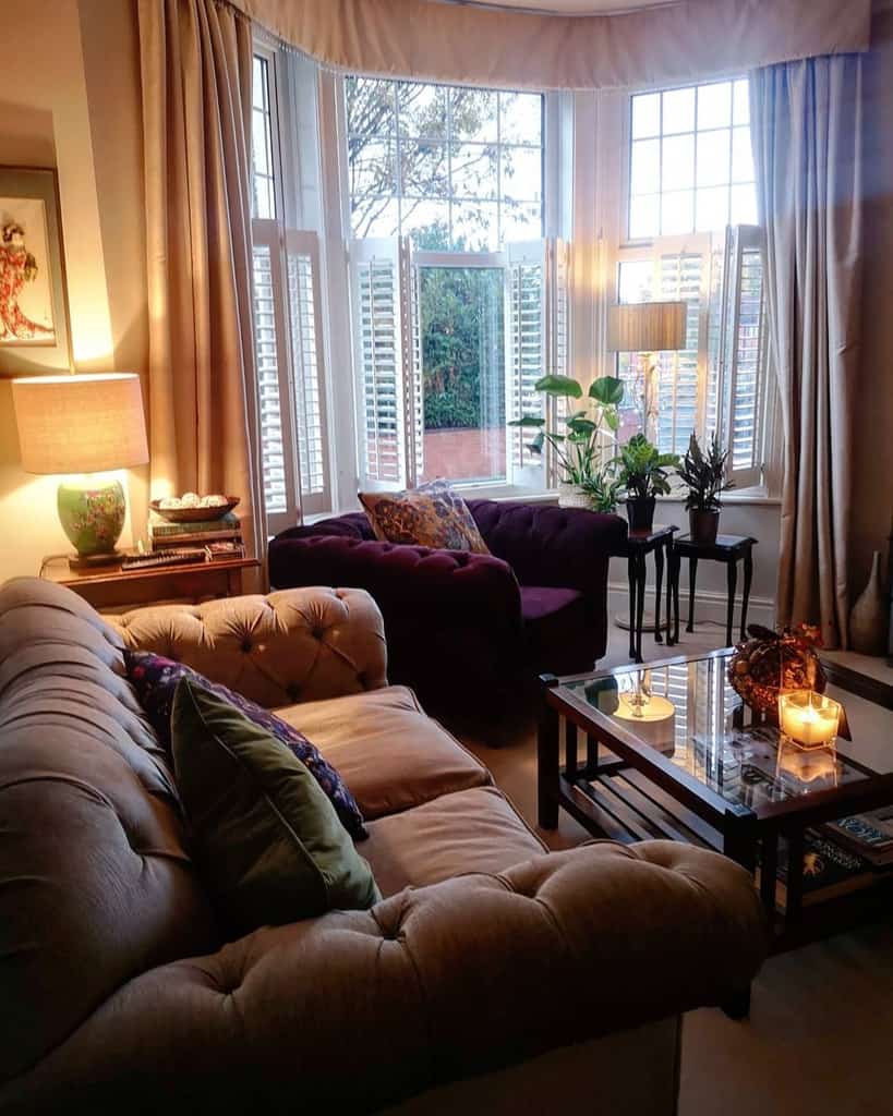 Elegant bay window living room with plush tufted sofas, soft drapes, warm lighting, and cozy candles, creating an inviting and classic ambiance.