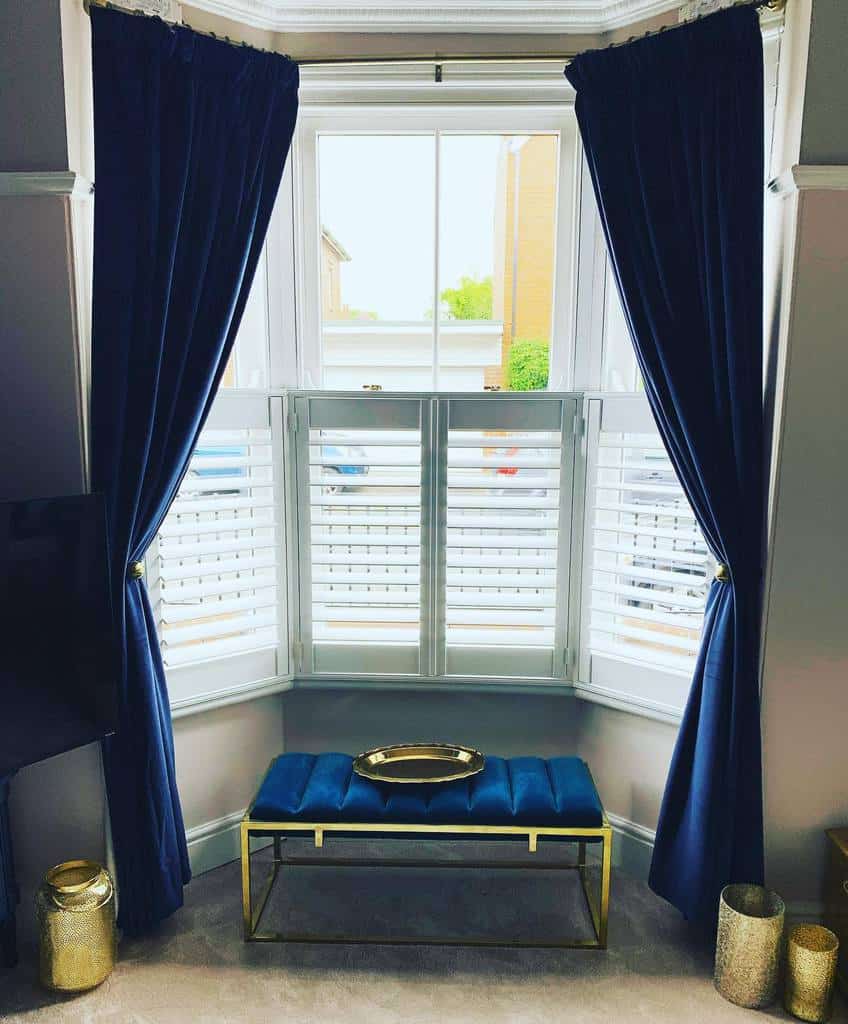 Sophisticated bay window with navy velvet curtains, white plantation shutters, and a gold-accented bench, creating a luxurious and stylish space.