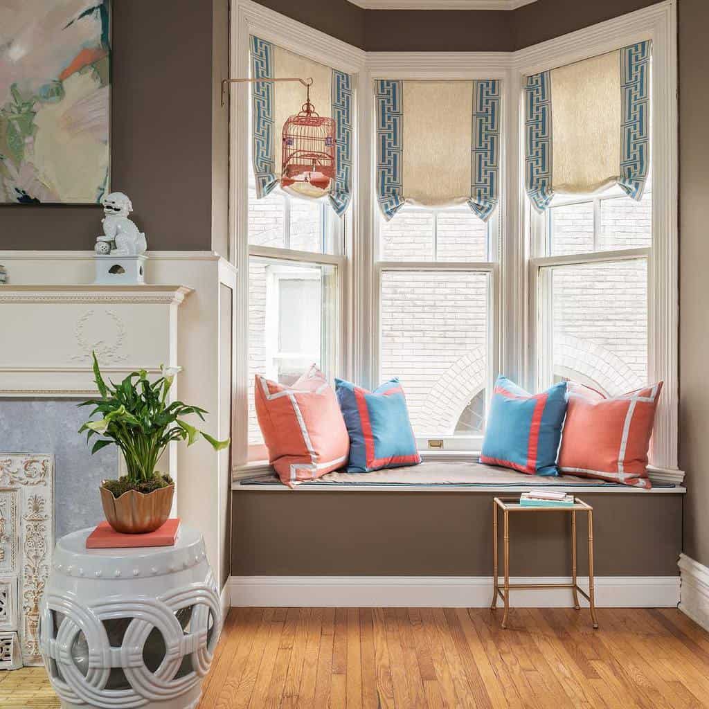 Charming bay window nook with a built-in bench, colorful pillows, patterned Roman shades, and classic decor, creating a cozy and stylish space.